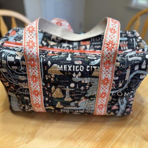 May include: A black travel bag with a map design featuring city names like Mexico City and Amsterdam. The bag has cream-colored straps with a red and white floral pattern. The bag is sitting on a wooden table.