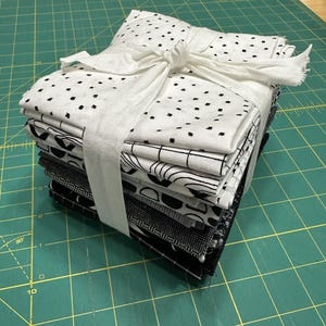 Peut inclure: Une pile de carrés de tissu pliés attachés avec un ruban blanc. Le tissu présente divers motifs noirs et blancs, notamment des petits points, des formes géométriques, des grilles et des textures noires unies, formant un ensemble monochrome.
