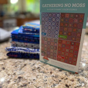 May include: A stack of folded blue and patterned fabric squares next to a rectangular instructional booklet titled GATHERING NO MOSS IN FOUR DYNAMIC COLOR STORIES, featuring four distinct quilt patterns in blue, green, brown, and red color palettes.