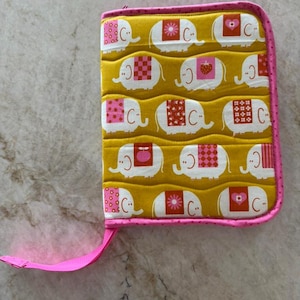 May include: A rectangular quilted fabric case featuring a mustard yellow background with a repeating pattern of white elephants. Each elephant has a unique pink or red patterned blanket on its back. The case has a bright pink zipper and trim.