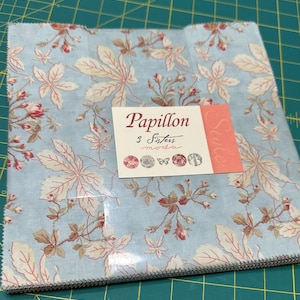 Może przedstawiać: Stos kwadratów materiału z kwiatowym wzorem w odcieniach niebieskiego, beżowego i czerwonego. Etykieta głosi "Papillon 3 Sisters moda" z etykietą "Style". Odpowiedni do pikowania lub innych projektów krawieckich.