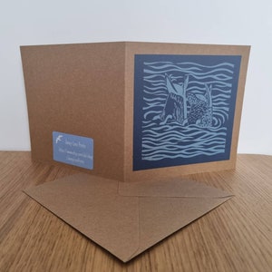 May include: A blue and white greeting card featuring a linocut design of two fish swimming in wavy water. The card is folded in half and comes with a brown envelope. The back of the card displays the text "Jacey Long Prints" and a website address.
