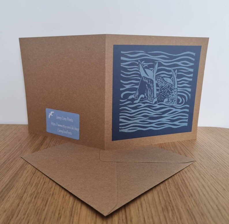 May include: A blue and white greeting card featuring a linocut design of two fish swimming in wavy water. The card is folded in half and comes with a brown envelope. The back of the card displays the text "Jacey Long Prints" and a website address.