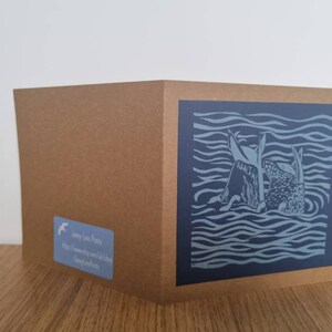 May include: A greeting card featuring a blue and white linocut print of two fish swimming in the sea. The card is on a brown background.