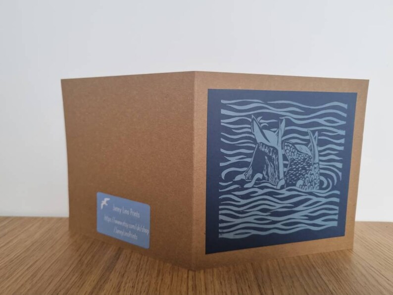 May include: A greeting card featuring a blue and white linocut print of two fish swimming in the sea. The card is on a brown background.