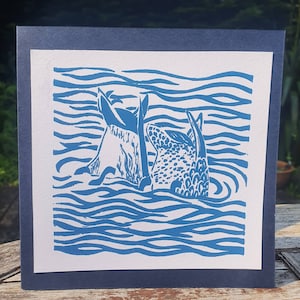 May include: A blue and white linocut print of two fish swimming in a wavy sea. The fish are facing each other and their tails are visible.