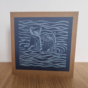 May include: A blue and white linocut print of two fish swimming in the ocean. The print is on a brown card.