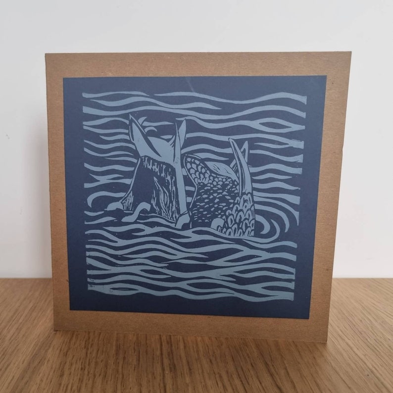 May include: A blue and white linocut print of two fish swimming in the ocean. The print is on a brown card.