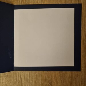 May include: A blank card with a white interior and a navy blue border. The card is folded in half and is lying on a wooden surface.