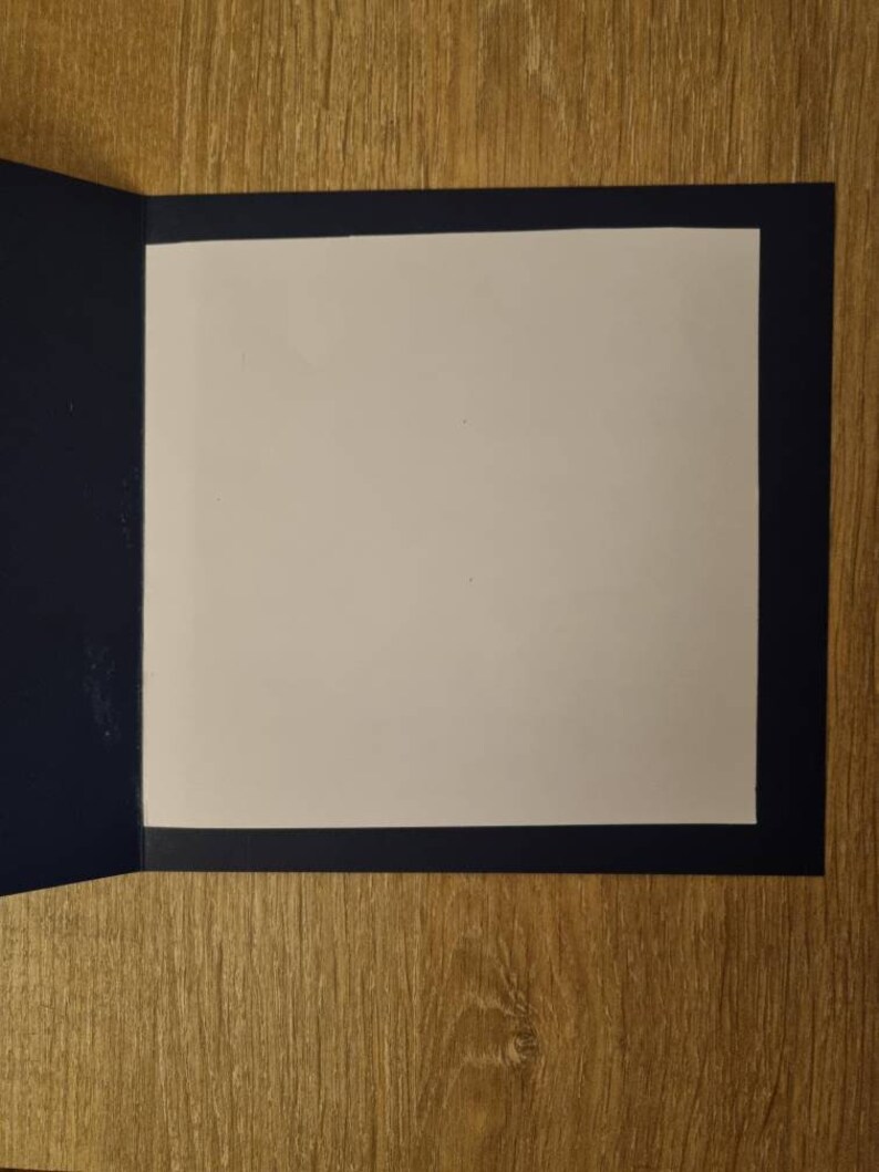 May include: A blank card with a white interior and a navy blue border. The card is folded in half and is lying on a wooden surface.