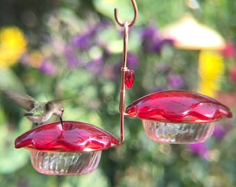 Op de afbeelding: Een rode glazen kolibrievoeder met twee voederstations. Een kolibrie drinkt nectar uit de voeder.