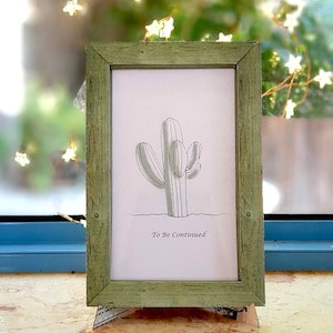 May include: A framed print with a green wooden frame. The print features a line drawing of a cactus with the text "To Be Continued" underneath.