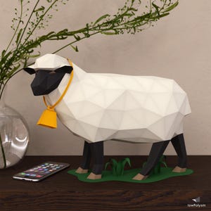 Puede incluir: Una oveja de papel blanco con patas negras y cara negra. La oveja tiene una campana amarilla alrededor del cuello y está de pie sobre una base de hierba verde.