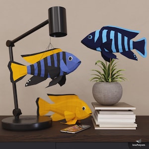 May include: A decorative arrangement featuring three colorful paper fish sculptures. One is suspended from a black lamp, while the others rest on a surface. The fish are blue, yellow, and black, with geometric designs. A small plant in a gray pot and a stack of books are also present.