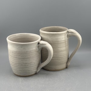 May include: Two white ceramic mugs with a textured, striped design. The mugs have a rounded shape and a handle on the side.