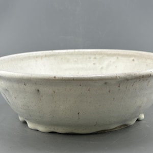 May include: A wide, shallow ceramic bowl with a speckled off-white glaze. The bowl features a rounded rim, a slightly uneven, hand-formed texture, and a base with small, integrated feet that elevate the vessel slightly from the surface.