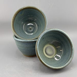 May include: Three handmade ceramic bowls in a stack. The bowls are a muted teal color with a light brown rim. The interior of the bowls have a glossy finish. The bowls are likely used for serving food.