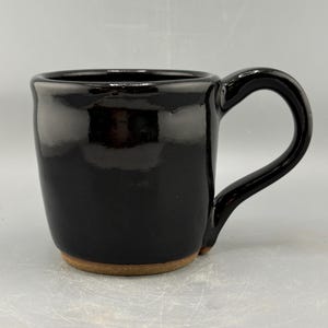 May include: A glossy black ceramic mug with a rounded body and a curved handle. The bottom of the mug has a natural, unglazed brown color. The mug is designed for hot beverages.