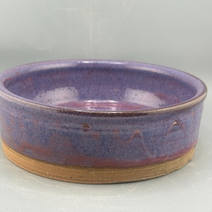 May include: A handmade ceramic bowl with a round shape. The bowl features a purple glaze with red undertones on the inside and exterior. The base is a natural tan color. This decorative bowl is suitable for serving food or as a decorative piece.