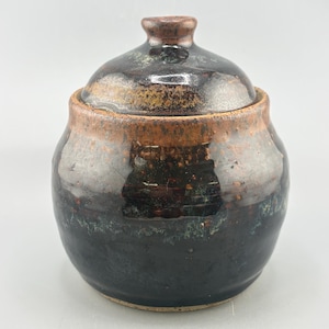 May include: A round ceramic sugar bowl with a matching lid, featuring a glossy dark brown and black glaze with mottled copper-toned accents around the rim and lid. The vessel has a wide, bulbous body and a small, knob-like handle on the lid.