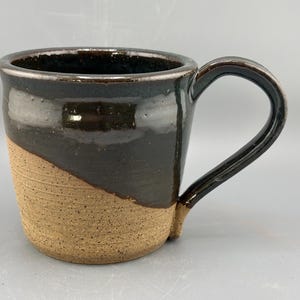 May include: A handmade ceramic mug with a dark brown glazed top and handle, and a tan, textured lower half. The mug has a curved handle and a slightly angled design, perfect for coffee or tea.
