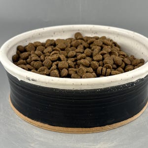 May include: A round ceramic bowl with a matte black exterior and a speckled white interior, filled with brown dry pet food kibble. The bowl features a wide rim and a visible unglazed clay base, showcasing a two-tone design with horizontal ridges.
