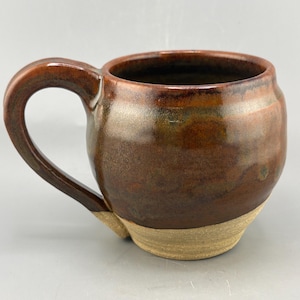 May include: A rounded ceramic mug featuring a glossy, deep brown and bronze glaze on the upper portion and a contrasting, unglazed matte beige base. The mug has a thick, curved handle attached to the side and a wide, circular opening at the top.
