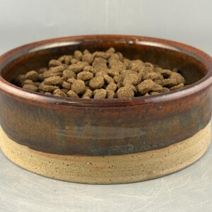 May include: A round ceramic pet bowl with a glossy dark brown glazed upper section and a textured, matte beige base. The bowl is filled with small, round, brown dry pet food kibble. The design features a distinct contrast between the smooth, dark rim and the earthy, speckled lower portion of the vessel.