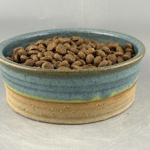 May include: A round ceramic bowl filled with brown dry pet food. The bowl features a two-tone glaze with a textured, speckled beige base and a smooth, deep blue upper section that transitions into a subtle green band where the two colors meet.