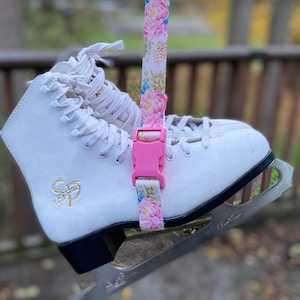 May include: White figure skates with a floral patterned strap and a pink buckle. The skates are hanging from a railing.