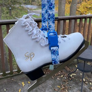 May include: White figure skates with a blue and white floral pattern strap. The strap has a blue buckle and is attached to the skate with a blue clip. The skates have a gold "SP" logo on the side.