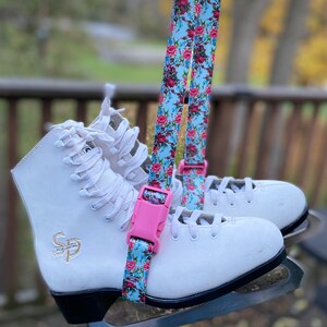 May include: A pair of white figure skates with black blades and a pink and blue floral skate strap with a pink buckle.