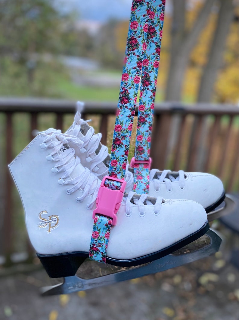 May include: A pair of white figure skates with black blades and a pink and blue floral skate strap with a pink buckle.