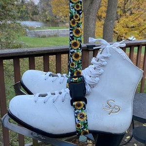 May include: A pair of white ice skates with black blades are hanging from a sunflower patterned strap. The strap has a black buckle. The skates have a gold logo on the side that says "SP".