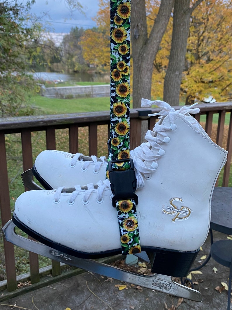 May include: A pair of white ice skates with black blades are hanging from a sunflower patterned strap. The strap has a black buckle. The skates have a gold logo on the side that says "SP".