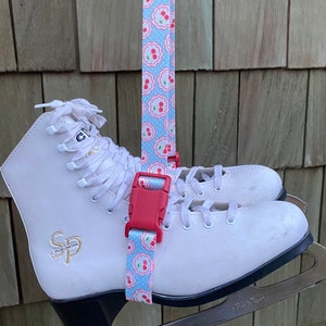 May include: White ice skates with a red and blue patterned strap attached to the top of the boot. The strap has a red buckle and the skates have a black base.