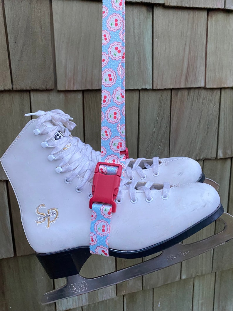 May include: White ice skates with a red and blue patterned strap attached to the top of the boot. The strap has a red buckle and the skates have a black base.