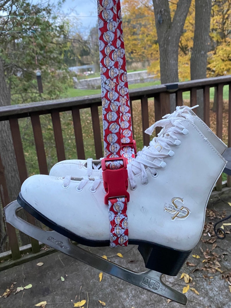 May include: White ice skates with a red and white Santa Claus pattern strap. The strap has a red plastic buckle. The skates have a black base and silver blades.