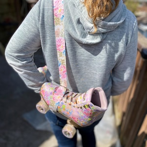 May include: A pink roller skate with a floral pattern is slung over the shoulder with a matching strap. The skate has a light pink boot, white laces, and clear wheels. The strap features pink roses and green leaves.
