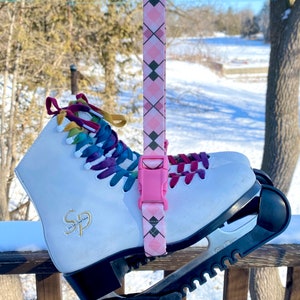 May include: White ice skates with a pink and black patterned strap attached. The skates have a black and white blade with a textured grip. The skates are on a wooden deck with snow in the background.