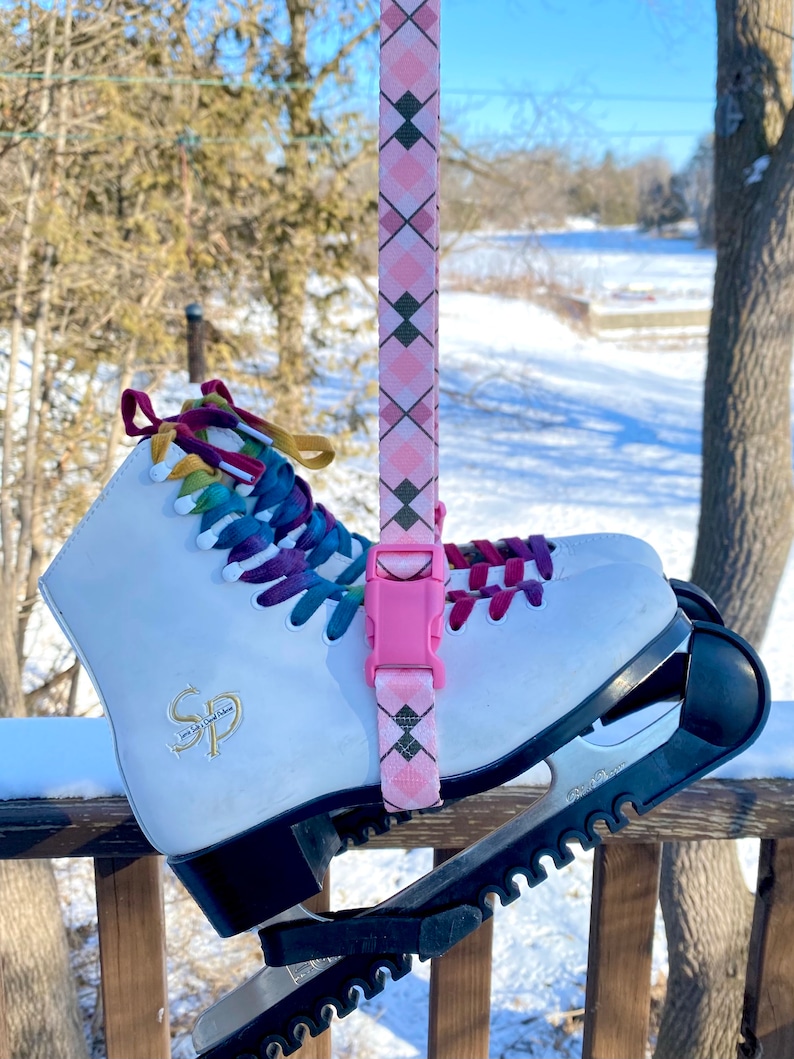 May include: White ice skates with a pink and black patterned strap attached. The skates have a black and white blade with a textured grip. The skates are on a wooden deck with snow in the background.