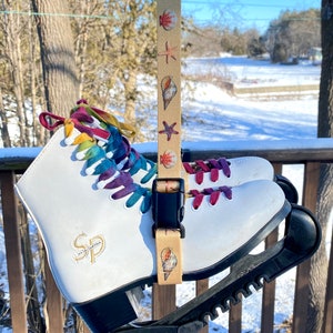 May include: White ice skates with rainbow laces and a black skate guard. The skates have a gold logo that says "SP" and are attached to a beige strap with a seashell pattern.