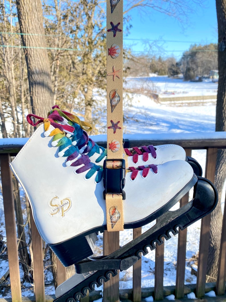 May include: White ice skates with rainbow laces and a black skate guard. The skates have a gold logo that says "SP" and are attached to a beige strap with a seashell pattern.