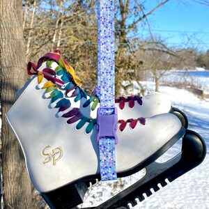 May include: White ice skates with colorful laces and a purple strap with a geometric pattern. The skates are attached to a black skate guard.