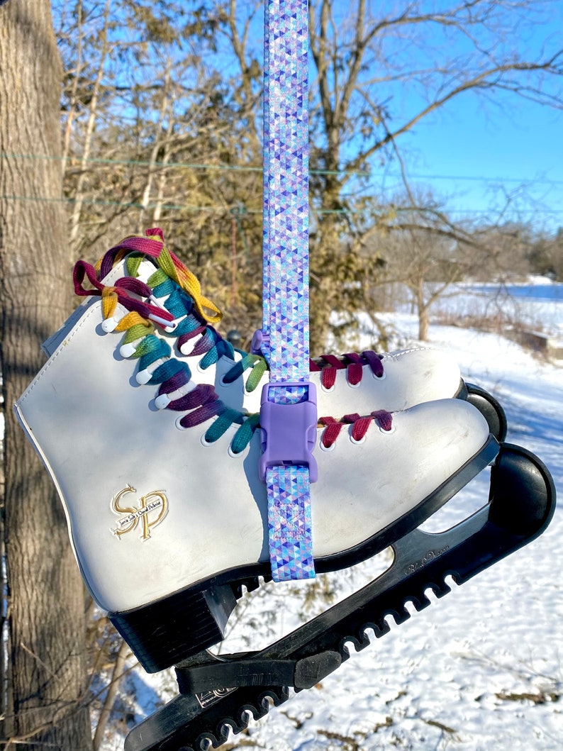 May include: White ice skates with colorful laces and a purple strap with a geometric pattern. The skates are attached to a black skate guard.