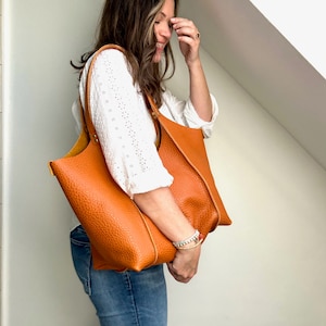 Op de afbeelding: Een vrouw in een wit, kant-topje en blauwe jeans houdt een grote, bruine leren tas met een bovenste handvat vast.
