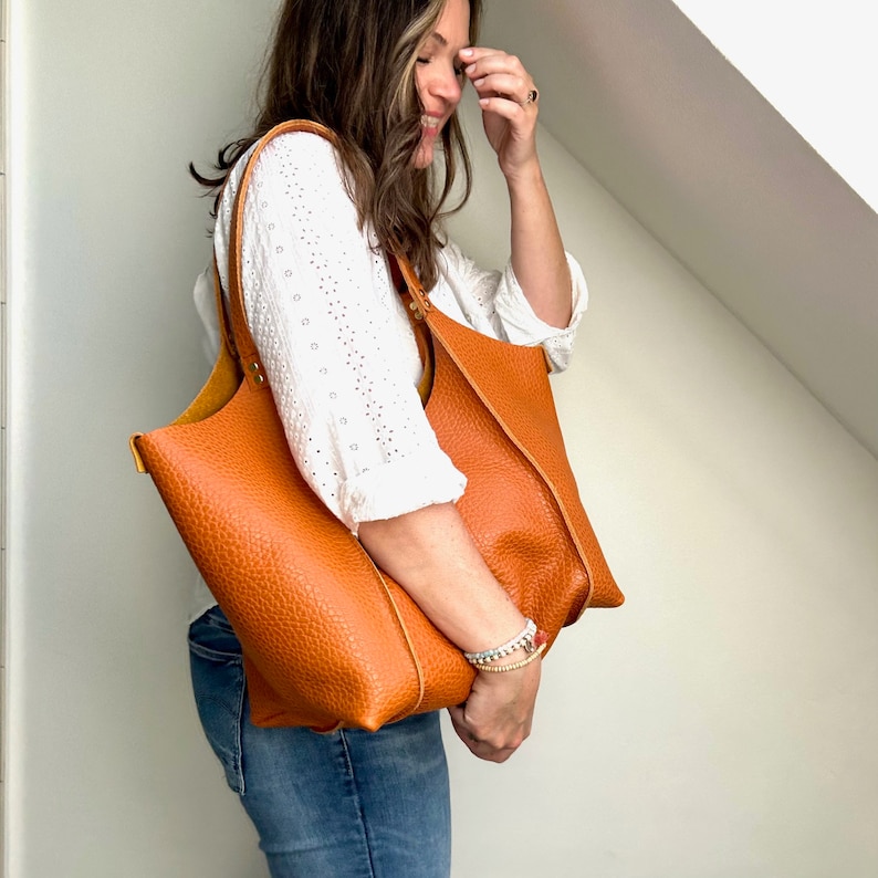Op de afbeelding: Een vrouw in een wit, kant-topje en blauwe jeans houdt een grote, bruine leren tas met een bovenste handvat vast.