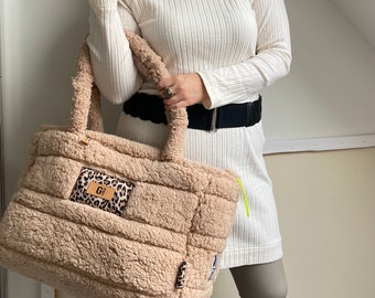 Op de afbeelding: Een grote, pluche, beige tote bag met een bruin leren patch waarop "G1" staat en een luipaardprint. De tas heeft een ritssluiting aan de bovenkant en wordt vastgehouden door iemand die een witte trui en een zwarte broek draagt.
