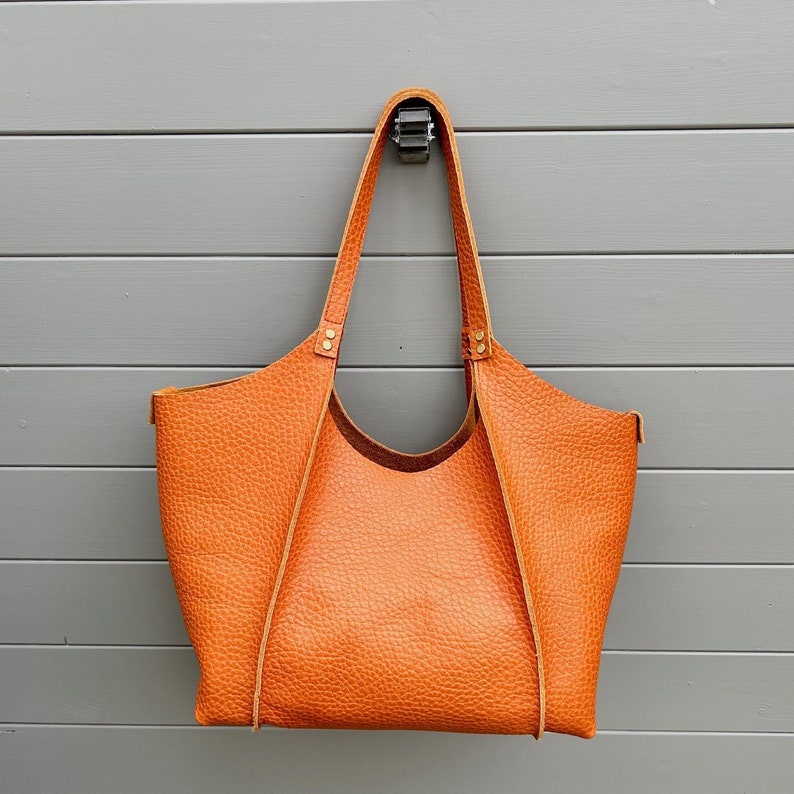 Op de afbeelding: Een grote, oranje leren tote bag met een afgeronde bodem en twee hengsels. De tas heeft een getextureerd, gegranuleerd oppervlak.