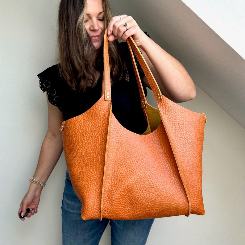 Op de afbeelding: Een grote, bruine leren tote bag met een gestructureerde, vierkante vorm. De tas heeft twee bovenste handvatten en een ritssluiting.